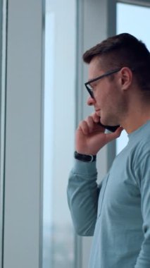 Beyaz bir erkek birini arıyor ya da telefona cevap veren birini dinliyor. Ofis penceresinde akıllı telefon kullanan bir iş adamı. Dikey video