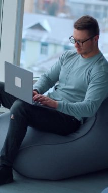 Genç adam rahat rahat koltukta oturuyor ve laptopuyla çalışıyor. Meşgul adam büyük pencerenin yanında çalışıyor. Yukarıdan bak. Dikey video