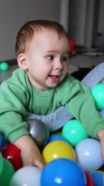 Mutlu gülümseyen çocuk kuru havuza yaslandı ve toplarla oynadı. İçeride oynayan sevimli bir bebek. Dikey video