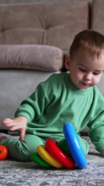 Küçük bebek halının üzerinde oturup oyuncak piramitle oynuyor. Çocuk oyuncağı çevirir ve bir yüzüğü ağzına çeker. Dikey video