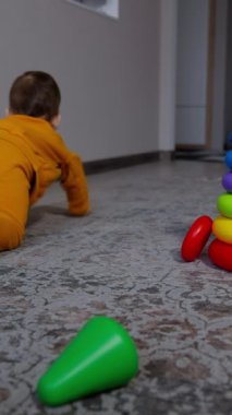 Bebek odada sürünüyor, parlak oyuncak piramidini görmezden geliyor. Turuncu takım elbiseli çocuk duvarın yanında duruyor. Dikey video