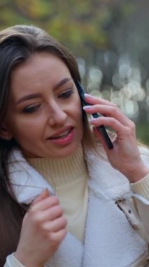 Dışarıda telefonda konuşan mutlu, güler yüzlü bayan. Çekici esmer, sonbahar parkında telefonundan selfie çekiyor. Arkaplan bulanık. Dikey video