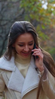 Beyaz bir kadın telefonda konuşmasına son veriyor ve selfie çekiyor. Gülümseyen esmer kadın sonbahar parkında yürüyor. Dikey video