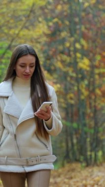 Yürüyen esmer, sonbahar parkında telefonuna odaklanmış. Uzun saçlı bayan geziniyor ve telefonunun ekranına bakıyor. Dikey video