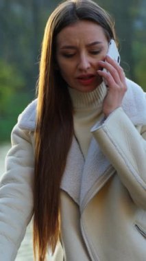 Açık havada telefonla konuşan esmer, ciddi bir bayan. Beyaz ceketli endişeli kadın nehir kenarında telefonla konuşuyor. Dikey video