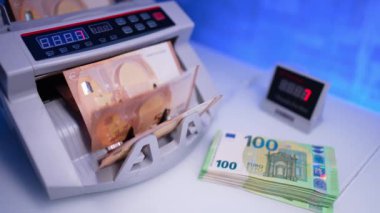 Çağdaş para sayma makinesinde dönen bıçaklarla çalışmak. Kadın eli, cihaza saymak için 50 euro veriyor. Masanın yanında 100 avroluk bir yığın duruyor ve yanında da 50 avroluk banknotlar bulunuyor. Kapat..