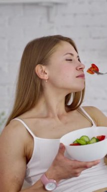 Salata yiyen genç bir kadın. Güzel bir kız mutfak arka planında organik sebzeleri kokluyor ve yiyor. Doğal gıda konsepti. Dikey video