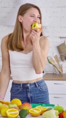 Kilo verme konsepti. Mutfakta meyve yiyen genç bir kadın. Organik vitaminlerle dolu bir masada duran kot pantolonlu güzel bir manken. Dikey video