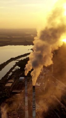 Gün batımında doğadaki enerji fabrikası. Akşamları nehir kenarındaki endüstriyel borulardan yoğun duman çıkıyor. Atmosferi kirletiyor. Hava görüntüsü. Dikey video