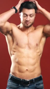 Gömleği olmayan yakışıklı sakallı adam. Kaslı vücutlu seksi bir sporcunun yarım boy portresi. Stüdyodaki koyu kırmızı arka planda kameraya bakan sağlıklı bir sporcu. Dikey video