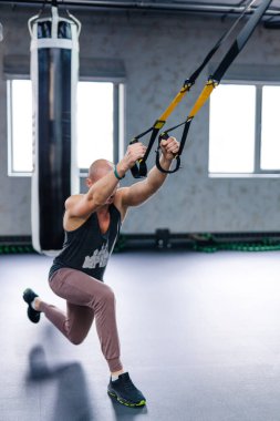 Adam modern spor salonunda TRX egzersizi yapıyor. Bir adam TRX kayışlarını gündüz saatlerinde parlak, ferah bir spor salonunda hareket egzersizi yapmak için kullanır..