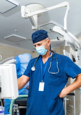 Cerrah hayati organlarını kontrol ediyor. Önlüklü bir tıp uzmanı hastane odasındaki ekipmanların izlenmesi ve hastanın güvenliğinin sağlanmasına odaklanır..