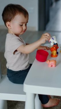 Güzel çocuk küçük masada oturup oyuncaklarla oynuyor. Bebek hayvanları ve renkleri inceliyor. Dikey video