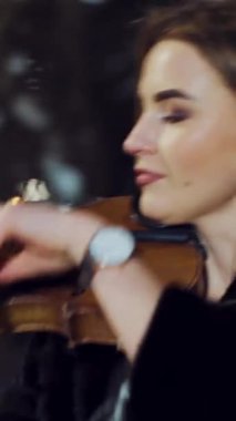 Çekici bir kız, akşam vakti sokakta siyah bir ceketle keman çalıyor. Dikey video etrafında kamera hareketi