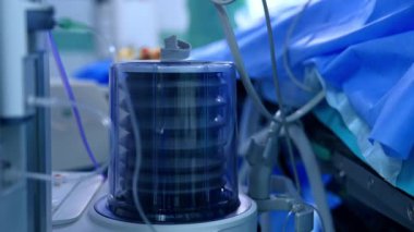 Yapay akciğer havalandırma makinesi pompası yavaşça yukarı ve aşağı gider. Mavi çarşaflı hasta yanında yatıyor..