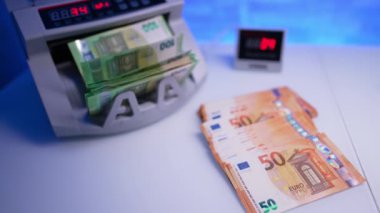 Masanın üzerinde portakallı 50 Euro var. 100 Euro 'luk banknotlar ortaya çıkıyor. Nakit sayma makinesi oluşuyor ve kadın eli onları masanın üstüne koyuyor. Kapat..