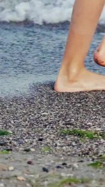 Dişi bacakları kapatın. Gün batımında sahilde yalınayak yürüyen bir kadın. Ağır çekim. Dikey video