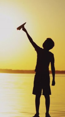 Mutlu çocuk kağıttan bir uçakla oynuyor. Nehirdeki ahşap bir köprüdeki çocuk gün batımında resim gibi bir ortamda. Dikey video