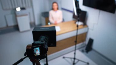 Stüdyoda günlük içeriği oluşturma. Kadın masada oturmuş, profesyonel kameraya bakıyor. Bulanık arkaplan.