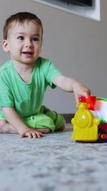 Gülümseyen beyaz çocuk yerde oturup oyuncaklarla oynuyor. Mutlu sağlıklı çocuk trenini zorluyor. Dikey video