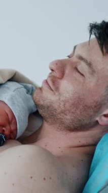 Koyu saçlı beyaz adam hastanedeki kanepede yatıyor. Baba, ağlayan yeni doğmuş bebeğini cilt teması için göğsünde tutuyor. Dikey video