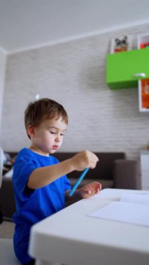 Lovely kid in blue t-shirt sits at desk holding a pencil. Baby boy starts drawing on the white paper sheet. Vertical video.