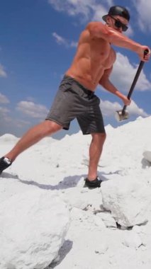 Doğada çalışan kaslı bir sporcu. Metal çekiçli güçlü bir adam dağda beyaz kayaları kırıyor. Dikey video