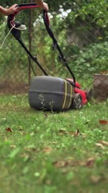 Bahçede elektrikli çim biçme makinesiyle çalışan bir kadın. Çim biçme makinesi dışarıda yeşil çim biçiyor. Dikey video