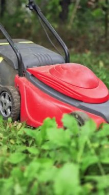 Bahçedeki kırmızı çim biçme makinesi. Çocuk dışarıda elektrikli çim biçme makinesi ile çim kesiyor. Dikey video