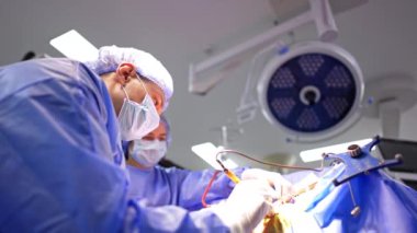 Konsantre erkek beyin cerrahı cerrahi bir cihaz kullanır. Ameliyat hemşiresi arka planda durup doktora yardım ediyor. Düşük açı görünümü.