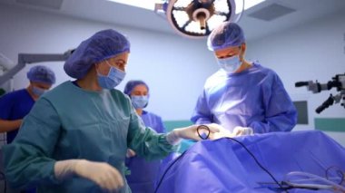 Kadın doktor tıbbi cihazın kablosunu çözüyor. Mavi cüppeli, lateks eldivenli, maske ve şapkalı kadın cerraha şırınga veriyor. Nöroşirürji için hazırlık safhası.