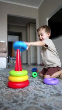 Tatlı erkek bebek yerde oturup piramit inşa ediyor. Aktif çocuk evde oyuncaklarla oynuyor. Dikey video.