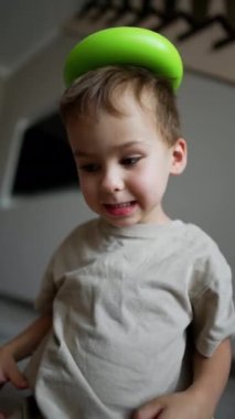 Komik çocuk evde neşeli bir şekilde oynuyor. Çocuk kafasına yeşil bir piramit takmış ve düşürmüş. Dikey video.