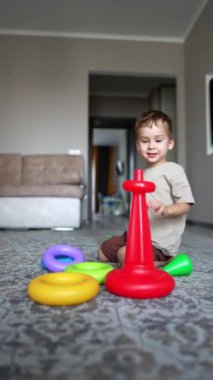Güzel gülümseyen beyaz çocuk evde neşeyle oynuyor. Küçük çocuk bir piramit inşa ediyor. Dikey video.