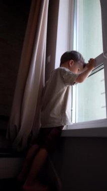 Tişört ve şort giyen beyaz çocuk pencerede duruyor. Küçük çocuk dışarı bakarken cama doğru eğildi. Düşük açı. Dikey video.