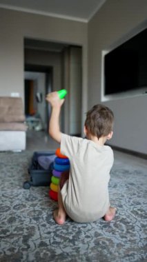 Oyuncak piramidin yanında oturan bir çocuğun arka görüntüsü. Toddler oyuncak detaylarını açık bavula koyar. Dikey video.