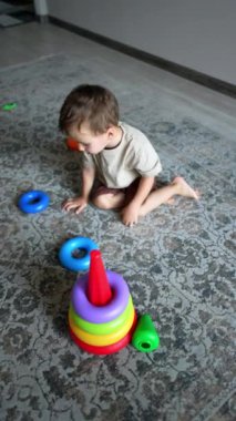 Küçük çocuk çabucak renkli bir oyuncak piramidi inşa etti. Çocuk yerde bir oyuncakla oynuyor. Üst Manzara. Dikey video.