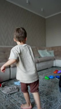 Sevimli çocuk odada dikilip arkasını dönüyor. Arka planda bazı oyuncaklar ve giysiler odanın etrafına saçılmış. Dikey video.