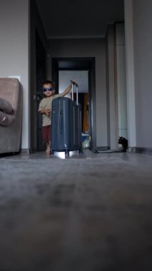 Güneş gözlüğü takan küçük sevimli çocuk odaya girip büyük bir bavul itiyor. Küçük bebek tatilci yolculuk için hazırlanıyor. Dikey video.