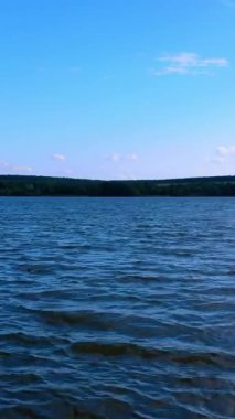 Gündüz vakti karanlık nehrin engebeli yüzeyi. İnsansız hava aracı alçaktan uçuyor ve sonra suyun üzerinde yükseliyor. Arka planda yeşil rıhtım var. Dikey video