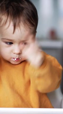 Bir yaşındaki çocuk yemek sırasında el sallıyor. Bebek, bir simit alırken annenin elini izliyor. Dudaklarında yemek olan sevimli bir çocuk. Dikey video