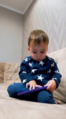 Mavi takım elbiseli şirin çocuk telefonla meşgul. Çocuk akıllı telefona odaklanmış bir şekilde kanepeye oturuyor ve parmağıyla kameraya dokunuyor. Düşük açı. Dikey video