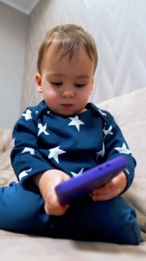 Kanepede sakince oturup telefonuna bakan güzel bir oğlan çocuğu. Sevimli çocuk kamerada dikkatini dağıtıyor ve parmağıyla dokunuyor. Düşük açı. Dikey video