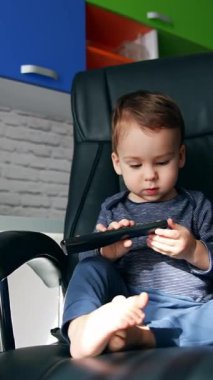 Meraklı çocuk elindeki TV kumandasına odaklanmış. Çocuk büyük deri koltukta oturuyor ve uzaktan kumandayı elinde tutuyor. Düşük açı. Dikey video