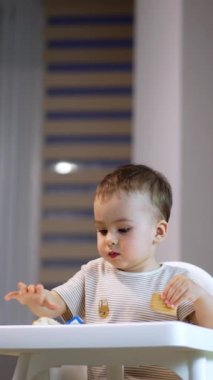 Yüksek sandalyedeki sevimli çocuk kurabiye ve çörek seçiyor. Bebek yemek için yemek seçiyor ve elinde bir çörek tutuyor. Dikey video