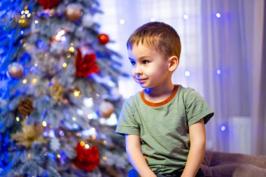 Mutlu çocuk tatil süslemelerinin tadını çıkarıyor. Neşeli bir çocuk güzel dekore edilmiş bir Noel ağacının yanında oturur, etrafı şenlikli ışıklar ve süslemelerle çevrilidir..