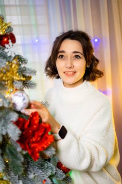 Kadın Noel ağacını neşeyle süslüyor. Bir kadın ışıldayan ışıklarla dolu bir odada Noel ağacına süs takarken gülümsüyor..