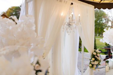 Çiçekli düğün zarafeti. Yumuşak beyaz perdeler, bir açık hava etkinliği sırasında çiçeklerle dolu büyüleyici bir düğün dekorunu ve narin bir avizeyi çerçeveler..