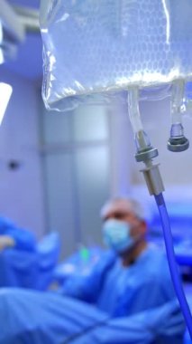 Salin solüsyonlu plastik torba ameliyathanede asılı. Arkaplanda hasta başında oturan bir erkek cerrahın bulanık görüntüsü. Dikey video.