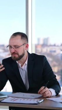 Sinirli iş adamı elinde telefonla masada oturuyor. Gözlüklü, akıllı telefon kullanan ve ofisinde kendi kendine konuşan orta yaşlı bir girişimci. İş sorunları. Dikey video